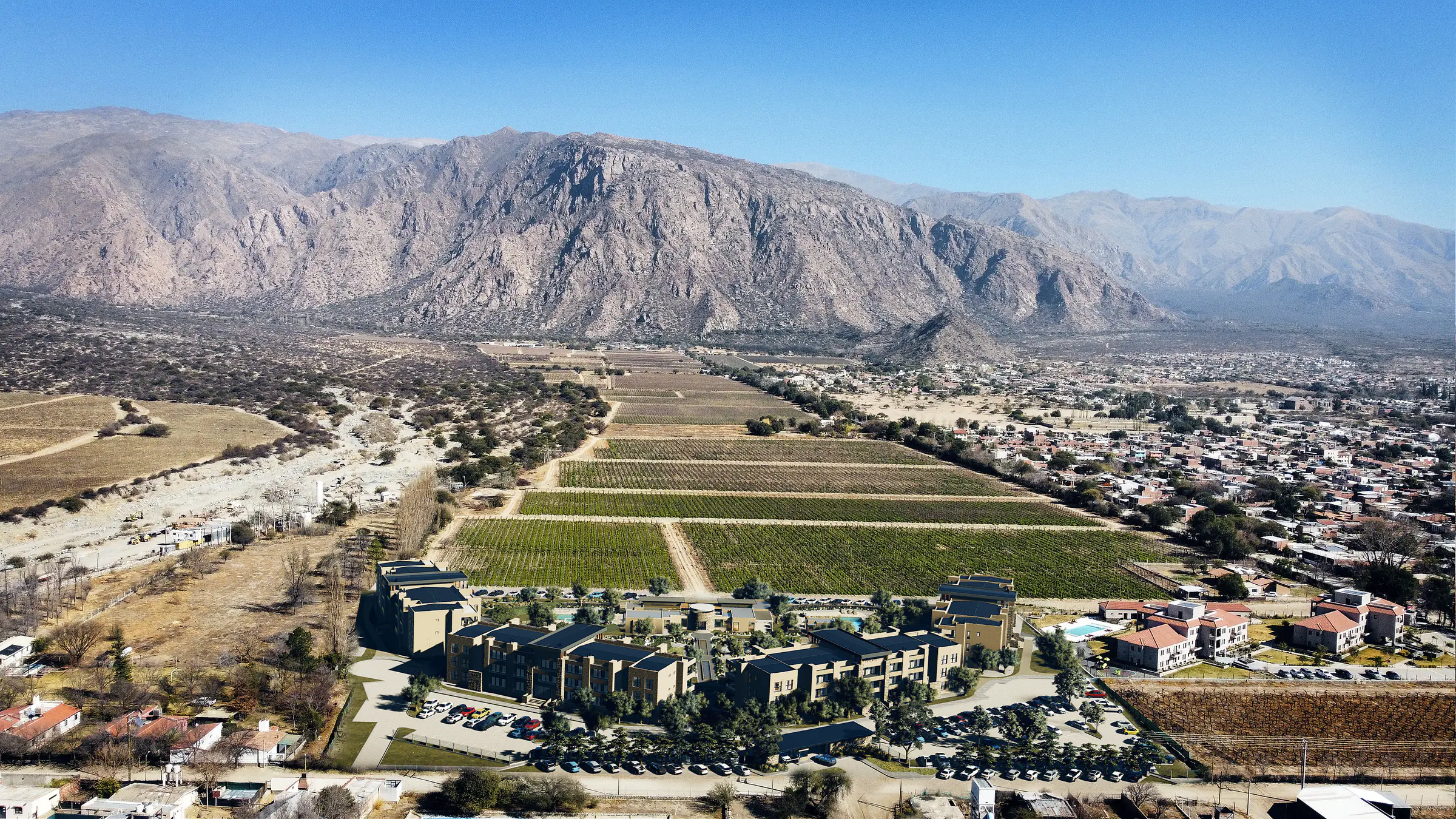 Vista aérea de día del complejo con los estacionamientos al frente; detrás, un viñedo grande y los cerros; a la derecha, las casas de Cafayate, y a la izquierda, el cauce seco del río