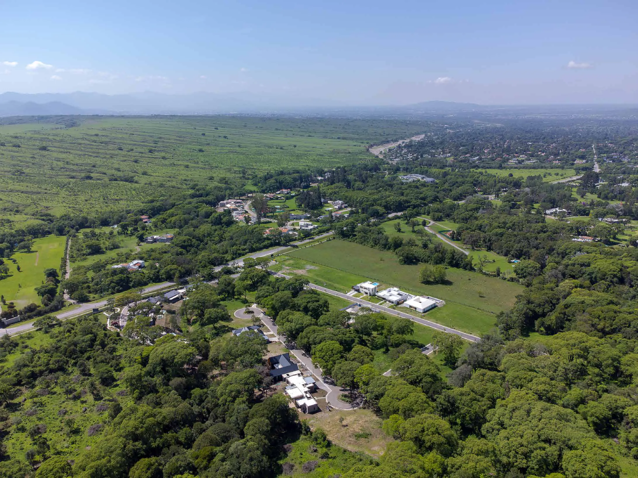 Vista aérea del barrio: una calle interna recta con tres casas blancas junto a un lote grande de pasto, una rotonda y casas entre árboles altos, y detrás el valle con otros barrios, campos y cerros en el horizonte
