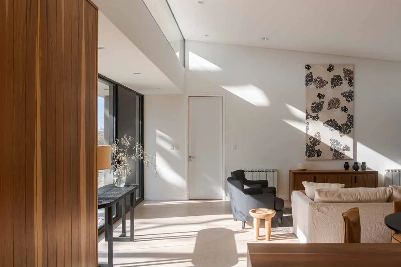 Living de una vivienda de El Retiro Housing: un sillón claro y dos butacas grises, una banqueta de madera, un aparador bajo un cuadro de manchas negras, un ventanal a la izquierda y un panel de madera en primer plano