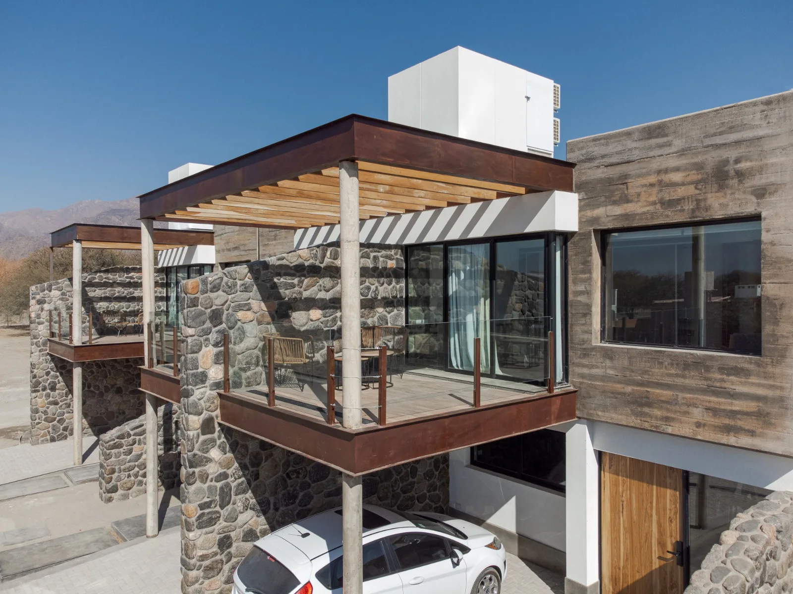 Una casa de muros de piedra y hormigón con textura de madera, con un balcón bajo una pérgola de madera y chapa oxidada, y un auto blanco en la cochera. Detrás, cerros bajo un cielo azul.