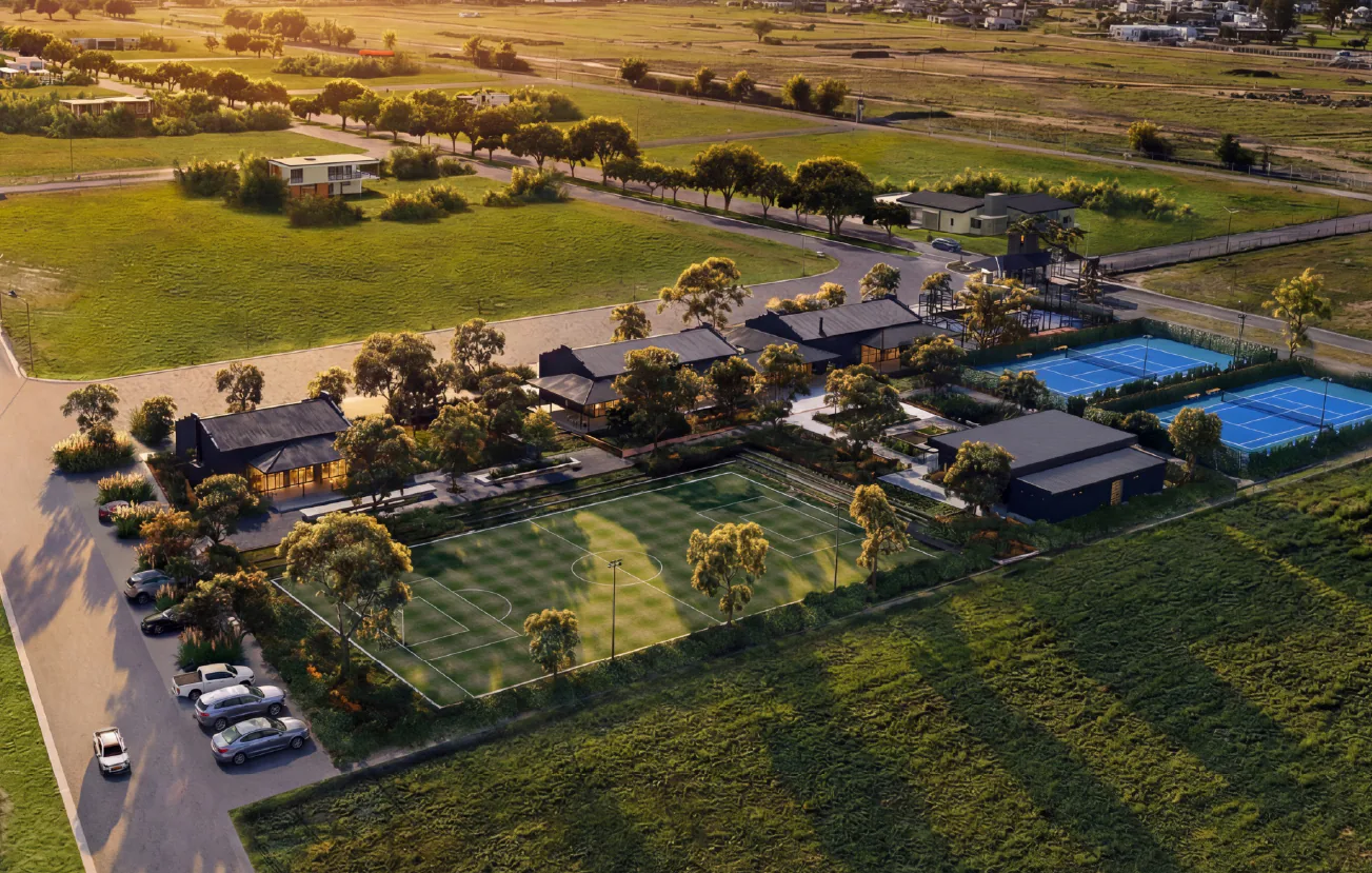 Render aéreo al atardecer del sector del club: edificios de techos oscuros entre árboles, una cancha de fútbol 7, dos canchas de tenis azules y un estacionamiento con autos, con campos y casas detrás.